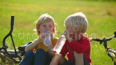 两个小男孩骑自行车的慢镜头坐在草坪上，从塑料瓶里喝水。 淡水概念